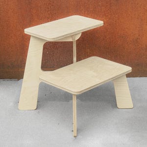 May include: A light-colored wooden desk with a two-tiered design. The top tier serves as a desktop, while the lower tier functions as a seat. The desk has a minimalist aesthetic, with clean lines and a natural wood finish. The desk is approximately 30 inches tall.