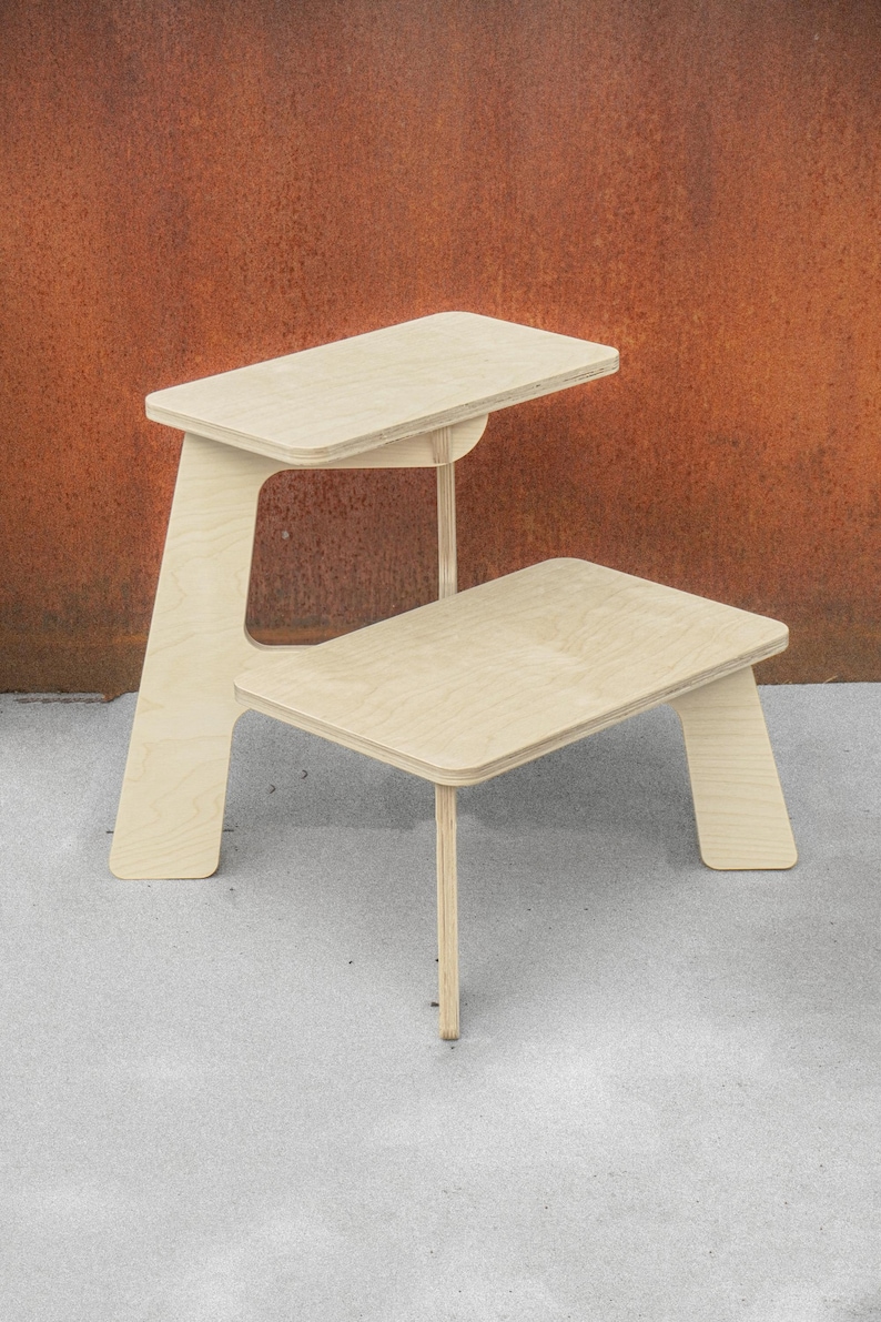 May include: A light-colored wooden desk with a two-tiered design. The top tier serves as a desktop, while the lower tier functions as a seat. The desk has a minimalist aesthetic, with clean lines and a natural wood finish. The desk is approximately 30 inches tall.