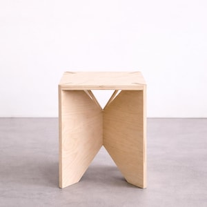 Puede incluir: Un taburete de madera clara con un diseño geométrico único. El taburete tiene una parte superior cuadrada y una base en forma de X. La veta de la madera es visible y el taburete está sobre un fondo neutro.