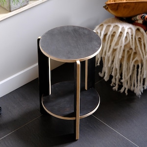 Puede incluir: Una mesa auxiliar moderna de dos niveles, con tapa y base redondas, y acabado negro. La mesa tiene un diámetro de aproximadamente 40 cm. El marco es de madera clara, creando un contraste con las superficies oscuras.