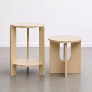 Peut inclure: Deux tables d'appoint en bois de couleur claire. La table la plus haute a un plateau rond, une étagère inférieure et quatre supports verticaux. La table la plus courte a un plateau rond et une découpe unique.