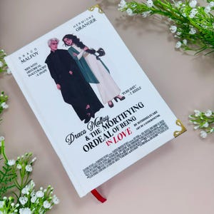 Könnte beinhalten: Ein weißes Buch mit dem Titel "Draco Malfoy & The Mortifying Ordeal of Being in Love", mit Illustrationen von Draco Malfoy und Hermine Granger. Es hat ein rotes Lesebändchen und goldene Eckdetails. Der Text enthält Zitate und Autoreninformationen.