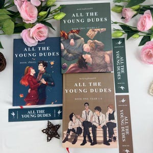 以下が含まれることがあります： 「All The Young Dudes」の書籍コレクション。表紙には若者のイラストが描かれています。本の色はネイビー、オリーブグリーン、タンなど様々です。同じタイトルのブックマークとピンクの花も一緒に写っています。