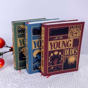 以下が含まれることがあります： 金色の文字で「All The Young Dudes」と書かれた3冊のハードカバー本。本は緑、青、バーガンディ色の表紙です。各表紙にはイラストと「All The Young Dudes」の文字が描かれています。