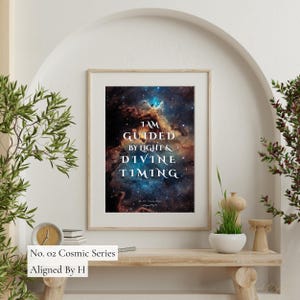 Puede incluir: Impresión artística enmarcada con una imagen vibrante de nebulosa y el texto "I AM GUIDED BY LIGHT & DIVINE TIMING". La impresión es parte de la "Cosmic Series" y está firmada "Aligned By H".