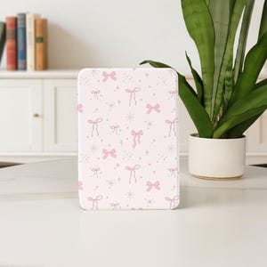 May include: A rectangular, pale pink object featuring a repeating pattern of pink bows, stars, and dots. The object is placed on a white surface, with a potted plant and a white cabinet in the background.