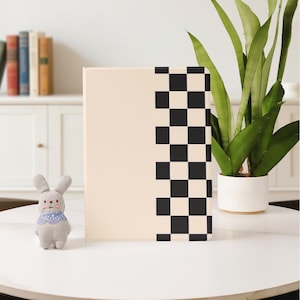 May include: A beige notebook with a black and white checkered pattern on the right side. A small gray bunny with a blue patterned collar sits to the left of the notebook on a white table. A green plant in a white pot is in the background.