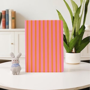 Op de afbeelding: Een notitieboek met verticale strepen in roze en oranje. Een klein grijs konijn met een blauw patroon zit links van het notitieboek. Een plant in een pot en een witte kast staan op de achtergrond. Het notitieboek is ongeveer 21 cm hoog.