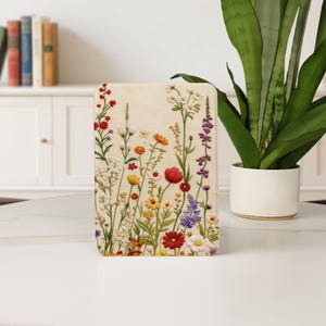 Puede incluir: Panel decorativo rectangular crema con un diseño floral bordado. El panel muestra una variedad de flores coloridas, incluyendo flores rojas, amarillas, moradas y blancas, con tallos y hojas verdes. Las flores están dispuestas en un patrón similar a un campo.