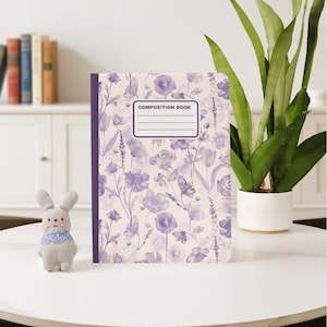 Peut inclure: Un cahier de composition avec un motif floral violet et blanc. La couverture porte le texte "COMPOSITION BOOK" dans une étiquette rectangulaire. Un petit jouet lapin gris avec un col bleu à motifs est à gauche du livre.