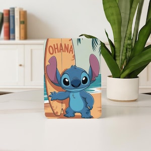 May include: A blue cartoon character with large ears stands in front of a surfboard with the word "OHANA" on it. The background depicts a beach scene with palm trees and ocean waves. The character is a popular animated figure.