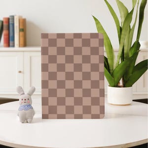 May include: A notebook with a tan and brown chequered pattern stands on a white table. A small stuffed bunny with a blue patterned collar is to the left of the notebook. A potted plant with long green leaves is to the right.