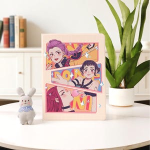 Puede incluir: Una funda beige para tableta con una colorida ilustración de estilo cómic de tres mujeres de estilo anime. La obra de arte incluye la palabra "DONE" en negrita. La funda está sobre una mesa blanca junto a un conejito de peluche y una planta en maceta.