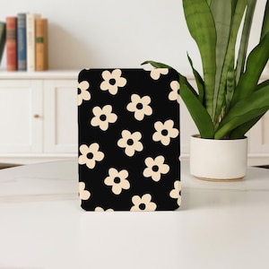 Peut inclure: Un étui rectangulaire noir avec un motif répété de fleurs de marguerite crème. L'étui est posé sur une surface blanche, avec une bibliothèque et une plante en pot en arrière-plan.