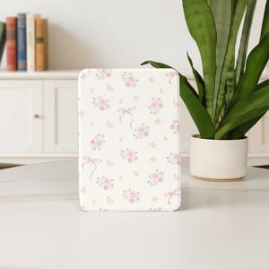 May include: A rectangular, cream-coloured item featuring a pattern of pink roses and bows. The design is repeated across the surface. The item is placed on a white surface, with a plant in a white pot to the right.