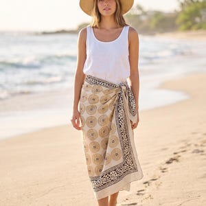 May include: A woman wearing a white tank top, a beige sarong with a circular pattern, and a wide-brimmed straw hat. The sarong is tied at the waist and has a black border. The woman is standing on a sandy beach near the ocean.