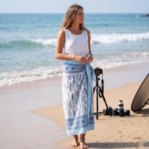 May include: A woman wearing a white tank top and a blue and white patterned sarong on a beach. The sarong features a repeating fish design and a striped border. The ocean and sand are visible in the background.