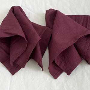 以下が含まれることがあります： 2つの折り畳まれたバーガンディ色の布ナプキン。ナプキンは質感のある生地でできており、白い表面に配置されています。ナプキンは、赤紫色の濃く豊かな色合いです。