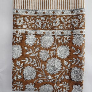 Könnte beinhalten: Gefalteter Stoff mit einem braunen und weißen Blumenmuster. Der Stoff zeigt ein sich wiederholendes Muster aus großen weißen Blumen und Blättern auf braunem Hintergrund. Der obere Rand hat einen gestreiften Rand.