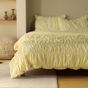 May include: A pale yellow bedspread and pillow set with a gathered design. The bedspread is on a bed frame, and the pillows are on top. The bedding is made of a soft, textured fabric. Decorative vases are on a shelf in the background.