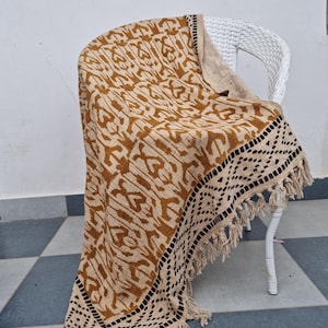 May include: A tan and brown patterned throw blanket with a black geometric border and fringe detail is draped over a white wicker chair. The blanket features a repeating abstract design.