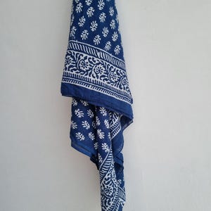 May include: An indigo blue textile with a white floral and leaf pattern, draped against a white background. The fabric has a detailed border design and a repeating pattern of small leaf-like shapes. The textile is hanging.