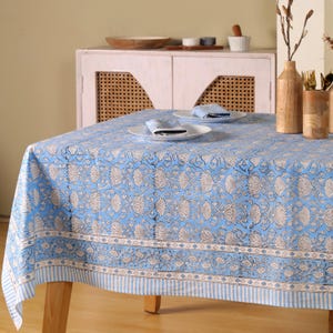 May include: A light blue tablecloth with a floral pattern in white and gray covers a wooden table. The tablecloth has a striped border and is set with white plates and napkins, creating a serene dining scene.