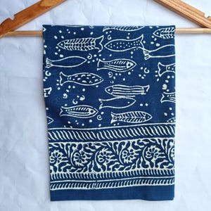 Peut inclure: Un tissu bleu marine avec un motif de poissons blancs et une bordure décorative. Le tissu est suspendu à un cintre en bois. Les poissons sont de styles variés et la bordure présente un motif floral.