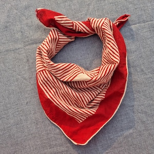 Può includere: Una bandana a righe rosse e bianche, piegata a triangolo. La bandana presenta un bordo rosso acceso e un motivo a righe bianche su sfondo rosso. Il tessuto sembra essere cotone o un materiale simile.
