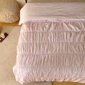 May include: A pale pink bedspread with a gathered, ruched texture. The bedspread covers a bed, with a matching pale pink sheet visible at the top. A cream-coloured cushion and a small rug are in the background.