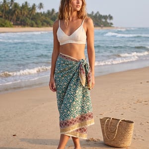 May include: A cream-colored bralette paired with a teal and cream patterned sarong featuring a dark red border. The sarong is tied at the waist. Also included are a woven straw tote bag and a pair of brown leather sandals on the sand.