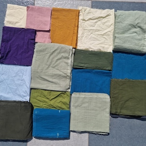 May include: A collection of colorful fabric squares in various shades, including purple, blue, green, and yellow. The fabric appears to be cotton or linen, with a slightly wrinkled texture. The squares are arranged on a tiled surface.