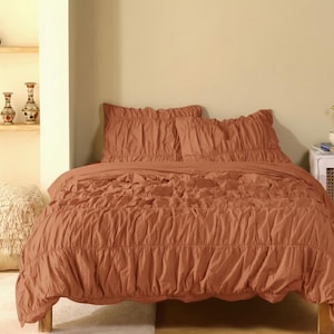 May include: A rust-colored bed set featuring a ruched design. The set includes a comforter and two pillow shams. The bedding is displayed on a wooden bed frame, creating a warm and inviting aesthetic.
