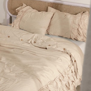 Può includere: Set di biancheria da letto beige con copripiumino e due federe. Il copripiumino ha bottoni in legno e un bordo a balze. Le federe hanno bordi a balze. La testiera è bianca con un design in canna intrecciata.