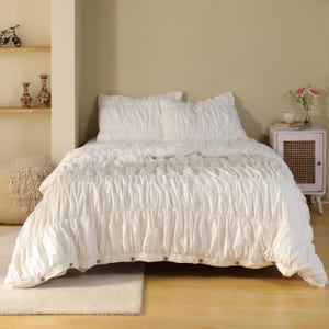 May include: A white bedspread and pillow set with a textured, gathered design. The bedding is on a bed with a wooden frame. The bed is in a room with light-colored walls and a wooden floor. The bedspread has wooden buttons.