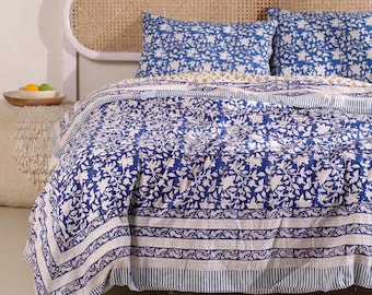 May include: A blue and white floral patterned quilt and matching pillowcases on a bed. The quilt features a repeating floral design with a border of stripes and decorative patterns. The headboard is a light color with a woven design.