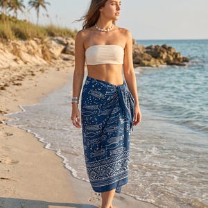 Peut inclure: Une femme porte un haut bandeau blanc et un sarong bleu avec un motif de poissons blancs. Le sarong est enroulé autour de la taille. Elle marche sur une plage de sable près de l'océan.