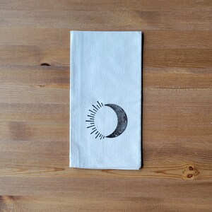 May include: A white tea towel with a black sun and moon design. The sun has rays and the moon has a crescent shape with small black dots.
