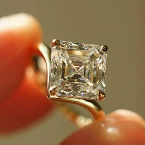 May include: A close-up of a gold ring featuring a large, square-cut diamond. The diamond is clear and multifaceted, reflecting light. The ring's band is a simple, polished gold. The ring is held between two fingers.