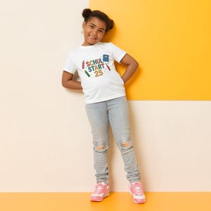 May include: A young person wearing a white t-shirt with the words "SCHUL START 25" printed on the front, along with colorful pencil and book graphics. The person is also wearing light blue jeans and pink sneakers.