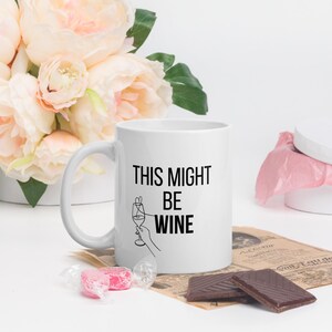 Puede incluir: Taza de cerámica blanca con el texto negro "THIS MIGHT BE WINE". La taza presenta un dibujo lineal de una mano sosteniendo una copa de vino. La taza está rodeada de flores, caramelos y chocolate.