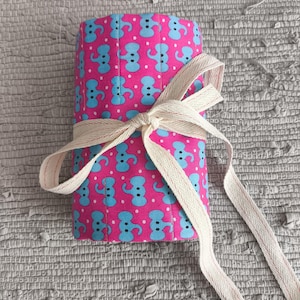 May include: A rolled-up fabric item with a pink background and a pattern of blue elephant shapes. A cream-colored ribbon is tied around the item in a bow. The item is resting on a textured gray surface.