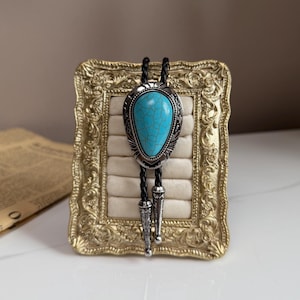 以下が含まれることがあります： シルバーカラーのメタルフレームと黒い革紐が付いた、ターコイズ色のボロタイ。ターコイズの石にはひび割れ模様があります。ボロタイは、金色の装飾フレームに飾られています。