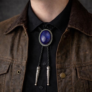 May include: A close-up of a bolo tie with a large, oval, deep blue stone set in a silver-colored metal frame. The tie has a braided black leather cord and decorative silver-colored tips. The person is wearing a black shirt and brown jacket.