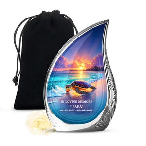 May include: A teardrop-shaped memorial urn with a beach scene featuring a sea turtle. The urn has a silver-colored trim and includes the text "IN LOVING MEMORY" and "PAPA" with dates. A black velvet bag and a white rose are also present.