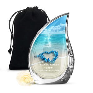 May include: A teardrop-shaped memorial urn with a beach scene and a heart made of blue butterflies. The urn has a silver-colored frame and a black velvet bag. Text on the urn reads "IN LOVING MEMORY 'PAPA' 01-16-1936 - 09-02-2018". A white rose is at the base.