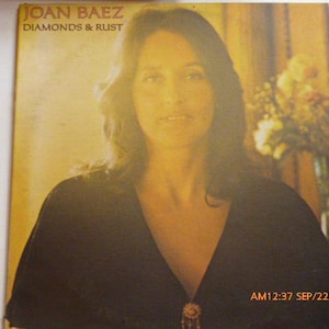 May include: A vinyl record cover featuring a portrait of Joan Baez with the text JOAN BAEZ DIAMONDS & RUST printed in the upper left corner. The image has a warm, golden-yellow tone, and the subject wears a dark top with a small decorative brooch.