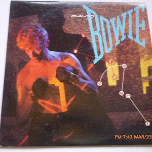 May include: Album cover featuring David Bowie with the word "BOWIE" in turquoise. The image shows a shirtless figure in a boxing pose, with a network of white lines and dots. The background is a mix of brown and yellow.