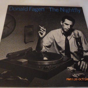 May include: Album cover for Donald Fagen's "The Nightfly" featuring a black and white image of a man in a shirt and tie, smoking a cigarette, near a record player and microphone. The album title and artist name are in blue text.