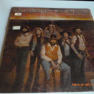 May include: A vinyl record album cover for The Charlie Daniels Band titled Million Mile Reflections. The cover features a group of five musicians standing and sitting on a stage in front of rows of empty theater seats, with a warm sunset background.